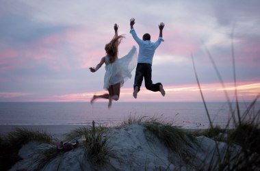 Ein Brautpaar springt bei untergehender Sonne glücklich von den Dünen, © Kinka Tadsen